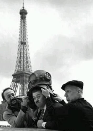 Photo en noir et blanc de Pierre Dac et Jean-Marie Amato en train de mettre un scafandre sur la tete de Francis Blanche, sur un pont devant la Tour Eiffel